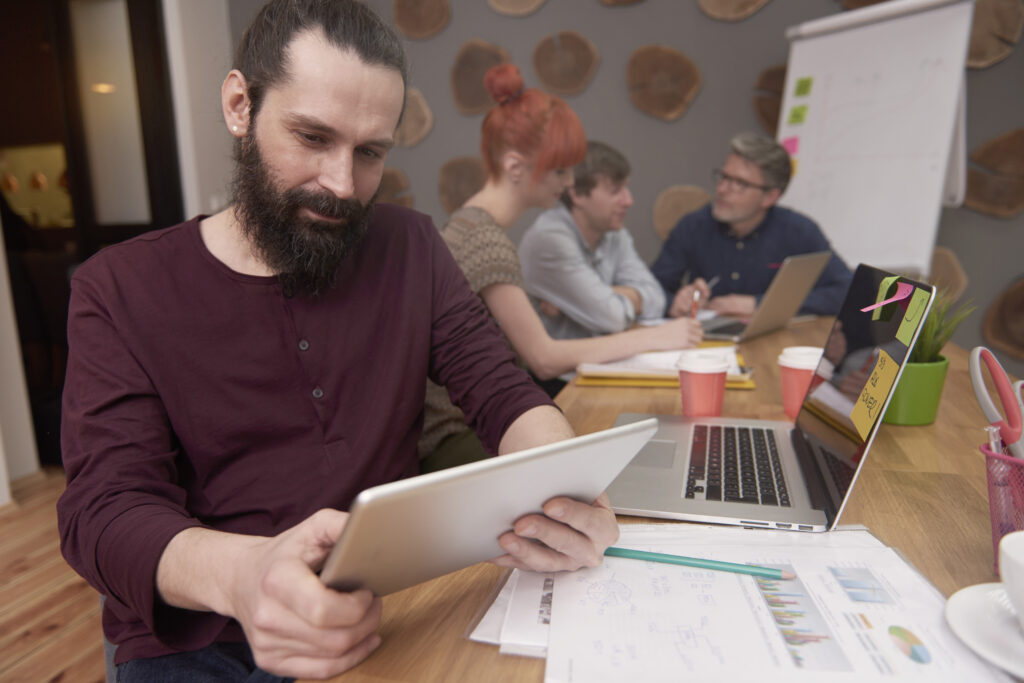 Eine Person arbeitet mit einem Tablet an einem Besprechungstisch, während weitere Personen im Hintergrund gemeinsam an einem Projekt arbeiten. Auf dem Tisch liegen Unterlagen, Diagramme und ein Laptop.