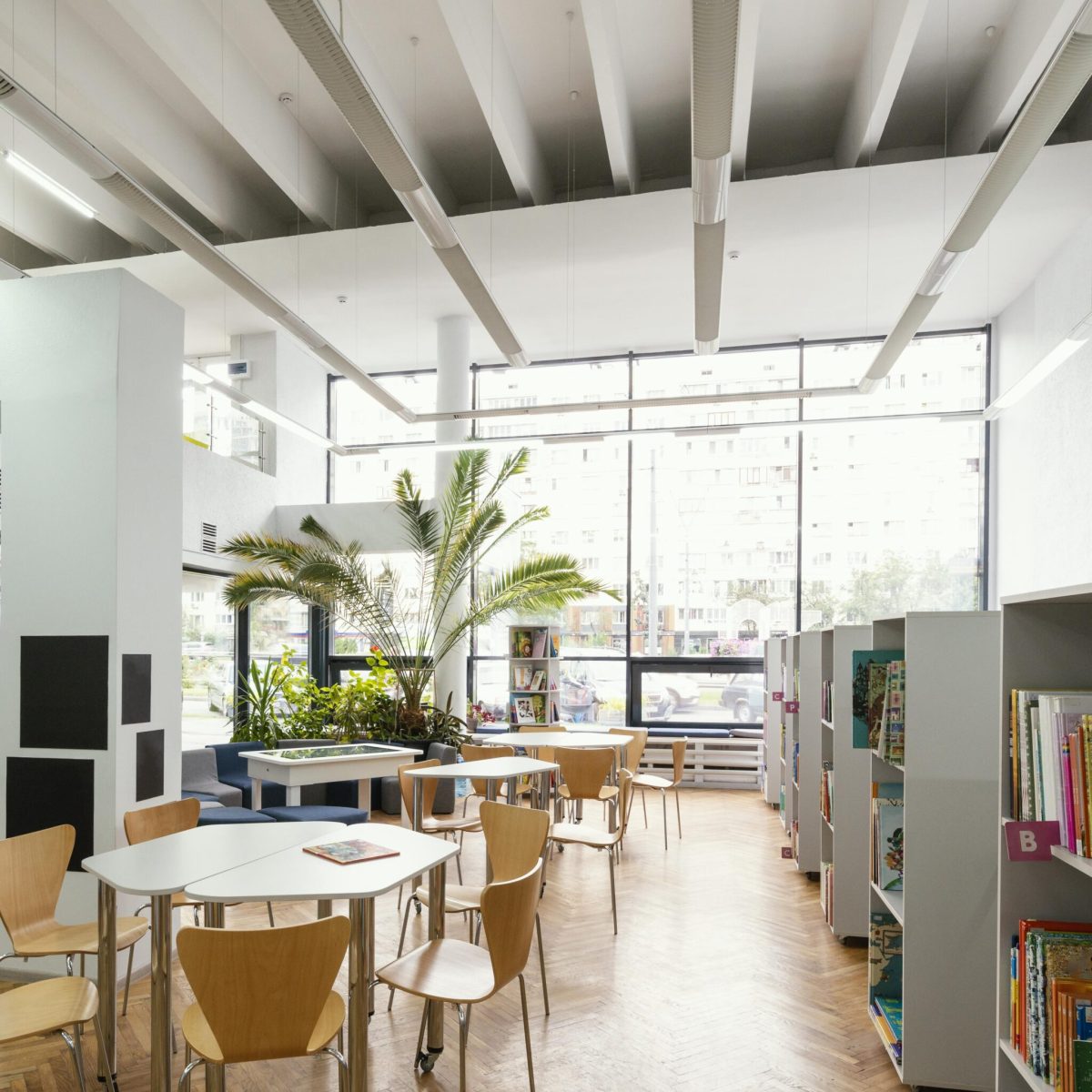 Heller moderner Lern- und Begegnungsraum mit Tischen, Bücherregalen und großen Fensterflächen.